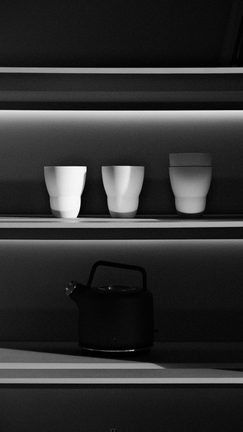 Three white cups and a black kettle on illuminated shelves.